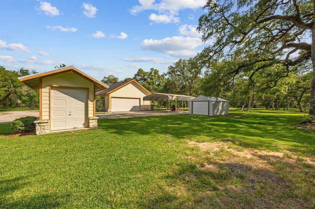 Cluster of functional outbuildings including the wellhouse, two car garage, covered carport and additional metal storage building—all situated on lush, open lawn under mature oaks.