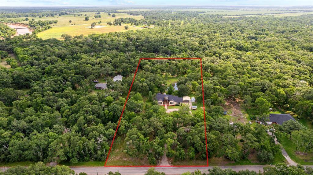 Top-down drone shot of the property: centered on the house’s roof, circular gravel drive, detached carport/shed, and pond just behind the home, all surrounded by mature trees.