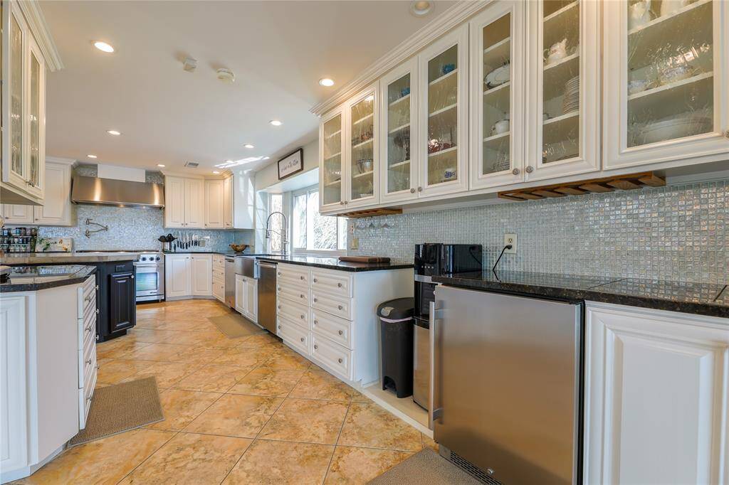 This kitchen features spacious granite countertops, white cabinetry with glass-front doors, and stainless steel appliances. The room is well-lit with recessed lighting and has a modern tile backsplash. A large window provides natural light, adding to the bright, open feel.