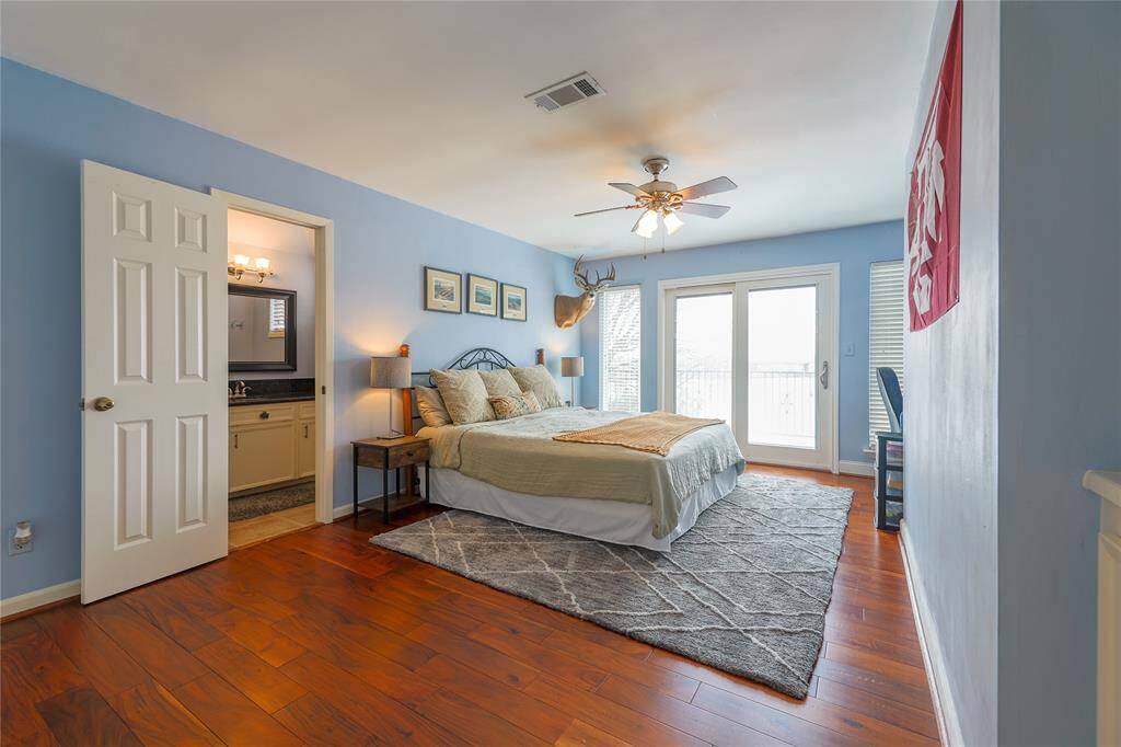 Bright and inviting bedroom with hardwood floors, soft blue walls, and a ceiling fan. It features ample natural light from large glass doors leading to a balcony, and an ensuite bathroom.