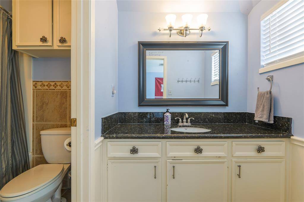 This bathroom features a classic design with a granite countertop, white cabinetry, and an elegant framed mirror. Light blue walls complement the neutral tile accents around the shower, creating a bright and welcoming space.
