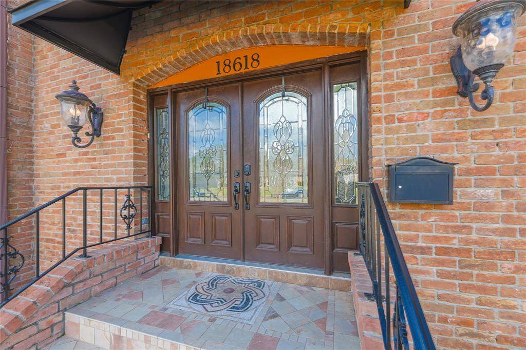This photo showcases an elegant brick entryway with a stately double-door featuring intricate glass designs. Flanked by ornate outdoor lanterns and complemented by a decorative tile step, it offers a welcoming and sophisticated entrance.