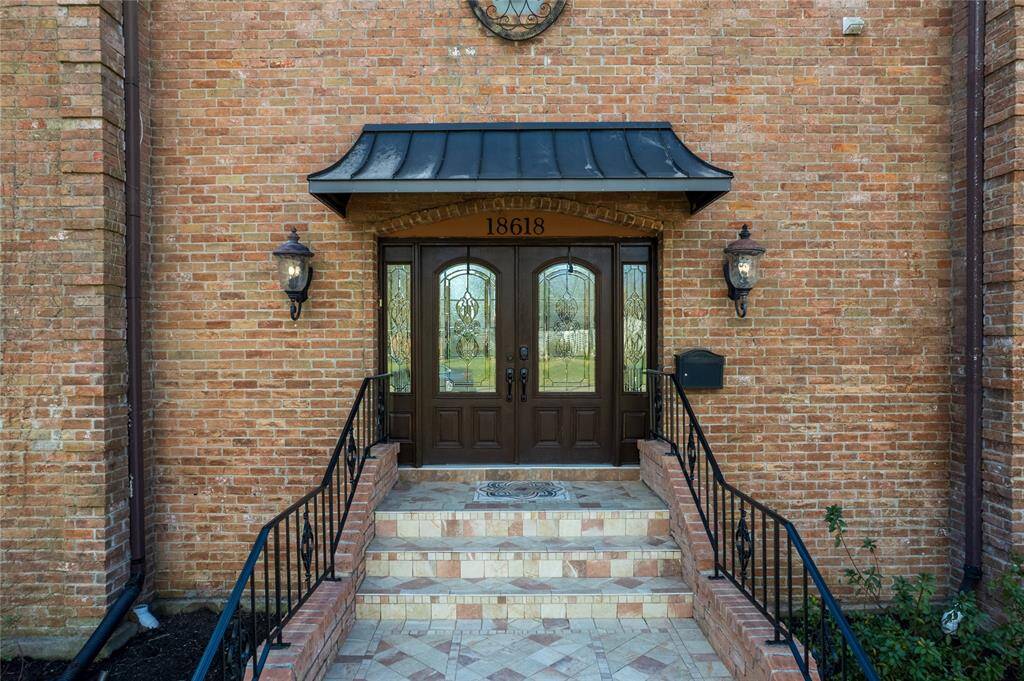 This photo showcases an elegant brick home entrance with a double front door featuring decorative glass panels. The entryway is framed by two vintage-style lanterns and a black metal awning. A short set of tiled steps, flanked by wrought iron railings, leads up to the doorway.