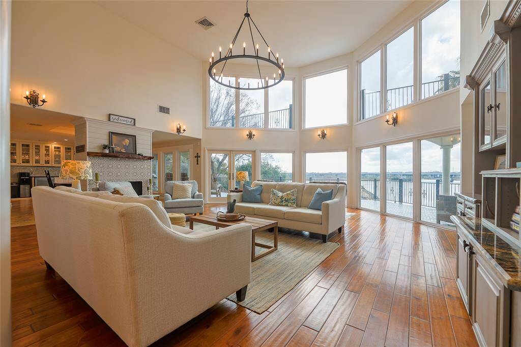 This spacious living room features high ceilings, large windows with water views, and abundant natural light. It includes elegant fixtures, a cozy fireplace, and built-in shelving, creating an inviting and stylish atmosphere perfect for relaxation and entertainment.