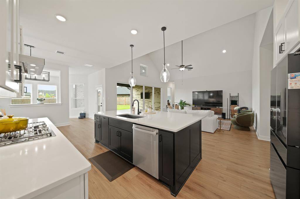 This photo shows a bright, open-concept living space with a modern kitchen featuring a large island, pendant lighting, and stainless steel appliances. The adjacent living area has high ceilings and a ceiling fan, offering a spacious and inviting atmosphere. Large windows provide ample natural light and a view of the backyard.