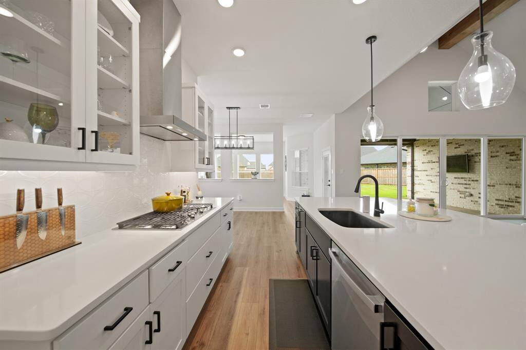 This modern kitchen features sleek white cabinetry, a spacious island with a farm sink, pendant lighting, and a view of the backyard through large windows. The open layout and wood flooring enhance the bright, airy feel.