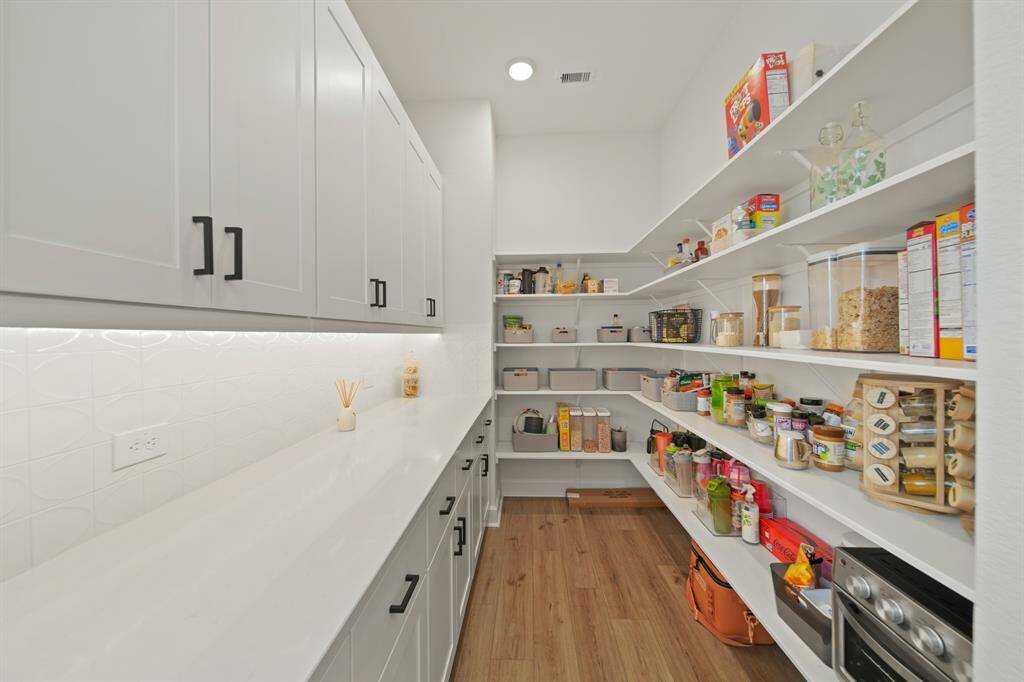 This photo showcases a spacious, modern pantry with ample shelving and cabinetry. The room features white cabinets with sleek black handles, under-cabinet lighting, and wooden flooring, creating a bright and functional storage space.
