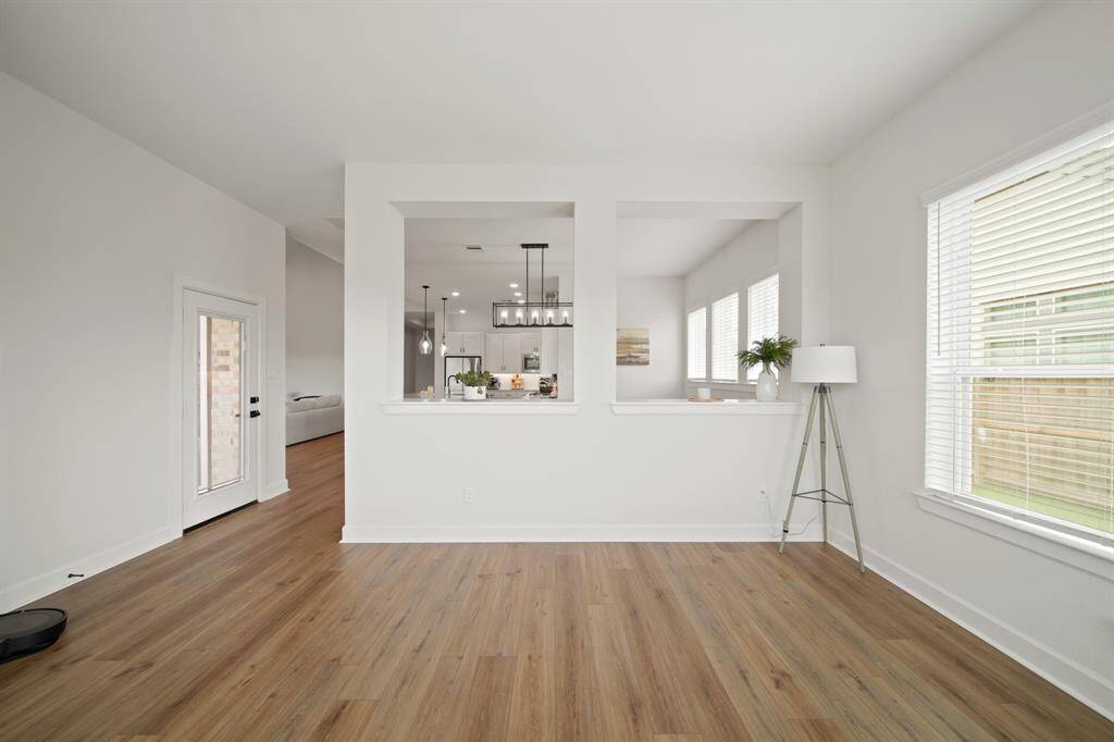 This photo showcases a bright, open-concept living area with wood flooring and large windows letting in natural light. The space features a modern kitchen visible through open wall cutouts, creating a seamless flow. A door leads to an outdoor area, enhancing the home's airy feel.