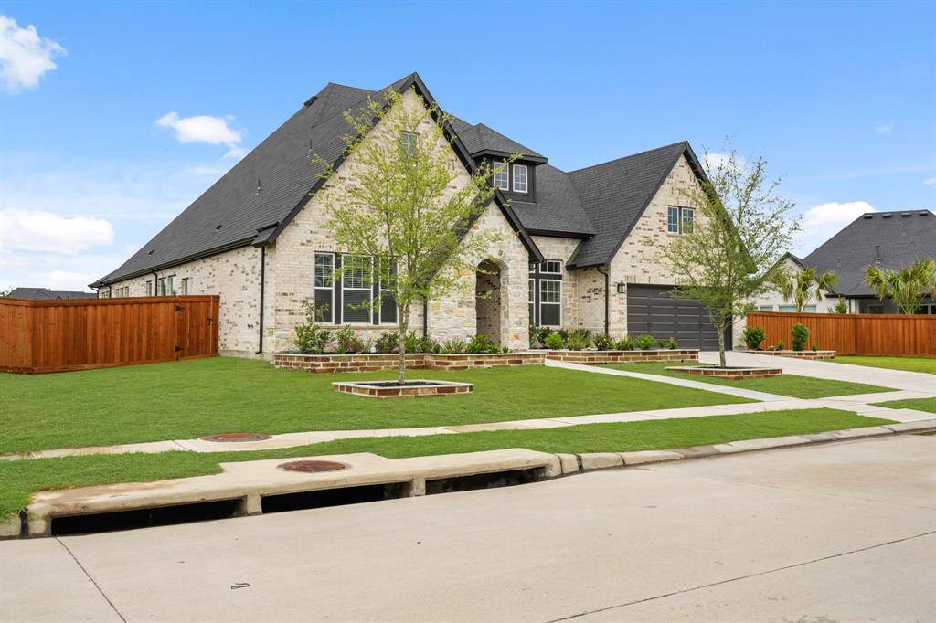 This is a modern,  brick home with a steep gable roof, featuring a well-manicured front lawn and a 4-car garage tandem. The property is surrounded by a wooden fence, offering a spacious driveway and attractive landscaping.