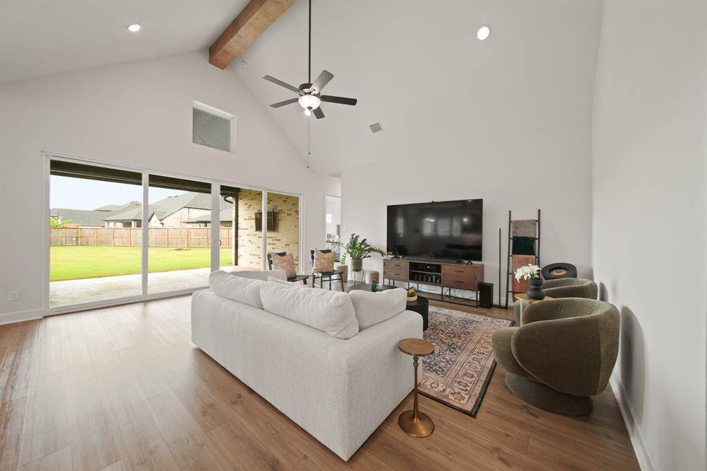 This spacious living room features high ceilings with a wooden beam, a large ceiling fan, and abundant natural light from sliding glass doors leading to a backyard. It includes a cozy seating area, modern furnishings, and a large TV, creating a perfect space for relaxation and entertainment.