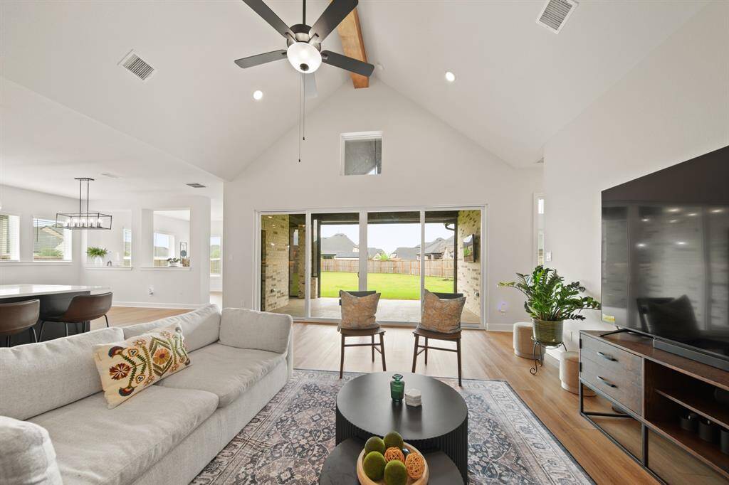 This spacious, open-concept living area features high vaulted ceilings, large sliding glass doors leading to a backyard, and abundant natural light. The modern design includes a cozy seating area, ceiling fan, and a seamless flow into the dining and kitchen spaces, perfect for entertaining.