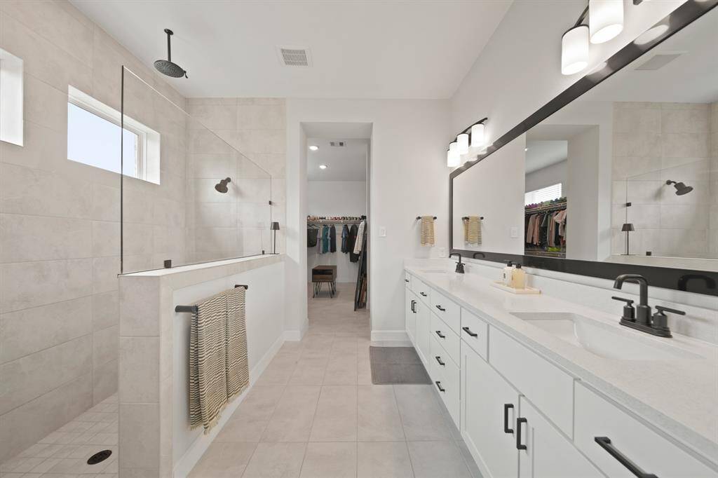 This spacious primary bathroom features a modern design with a large double vanity, sleek black fixtures, and ample storage. It includes a walk-in shower with elegant tile work and connects to a roomy walk-in closet.