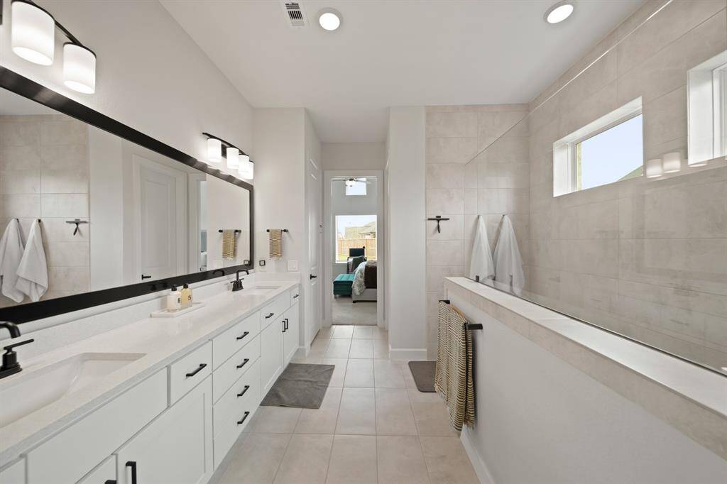 This spacious, modern primary bathroom features a long double vanity with sleek black fixtures and ample storage. Natural light filters in through well-placed windows, creating a bright and inviting atmosphere.