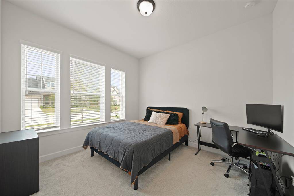 This bright bedroom features large windows, neutral carpeting, and a cozy layout with space for a bed and desk, ideal for comfort and productivity.