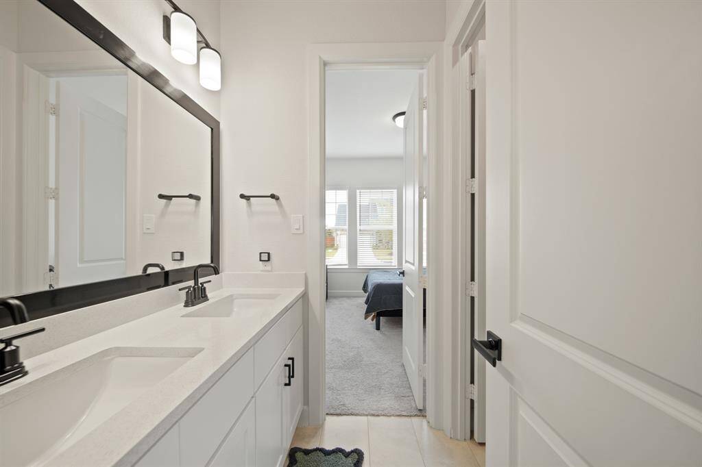 This photo shows a modern bathroom with a double vanity, sleek black fixtures, and a large mirror. It leads into a bright bedroom with carpeted flooring and a window providing natural light.
