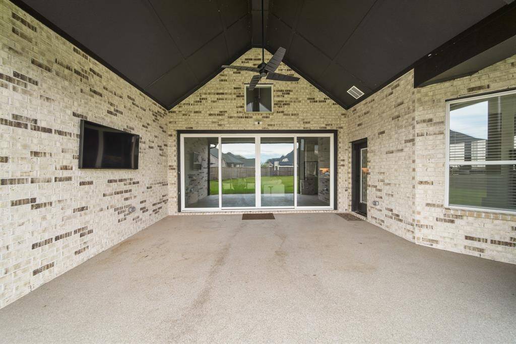 This photo showcases a spacious covered patio with brick walls, ceiling fan, mounted TV, and large sliding glass doors leading to a backyard, perfect for outdoor entertaining.