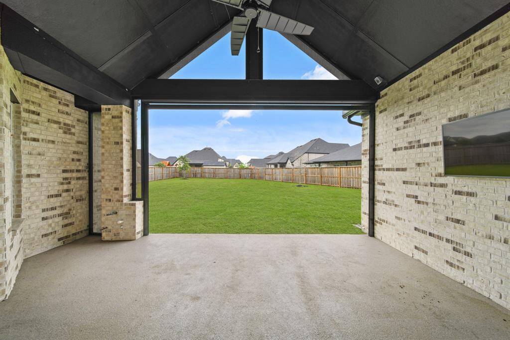 This photo showcases a spacious covered patio with a ceiling fan and mounted TV, overlooking a large, fenced backyard. The area features a vaulted ceiling, motorized screen and light brick walls, perfect for outdoor entertainment and relaxation.