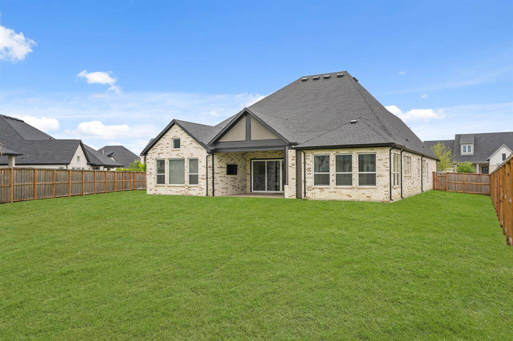 This photo showcases the backyard of a house with a spacious, well-maintained lawn and a covered patio area. The home features a light brick exterior with multiple large windows, surrounded by a wooden fence for privacy.
