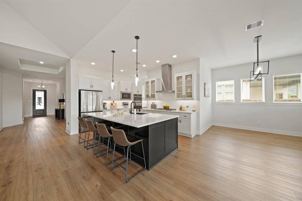 This open-concept kitchen features a spacious island with seating, modern pendant lighting, stainless steel appliances, and ample cabinet space. The room is bright with natural light from large windows and has stylish wood flooring throughout, creating a welcoming and functional space.