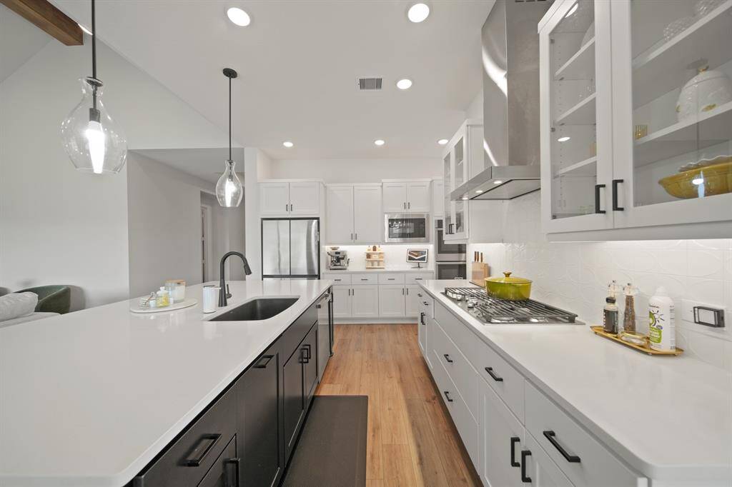 This modern kitchen features sleek white cabinetry, a large island with a sink and pendant lighting, stainless steel appliances, and wood flooring, offering a bright and inviting space for cooking and entertaining.