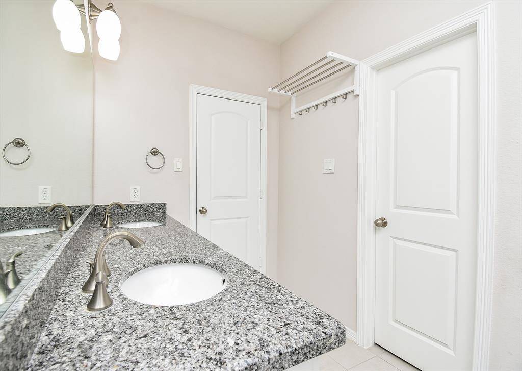 A spa-like setting where mornings begin in style: dual vanities, a generous soaking tub under a bright window, and a glass-enclosed shower—all framed by gleaming granite and soft white cabinetry.