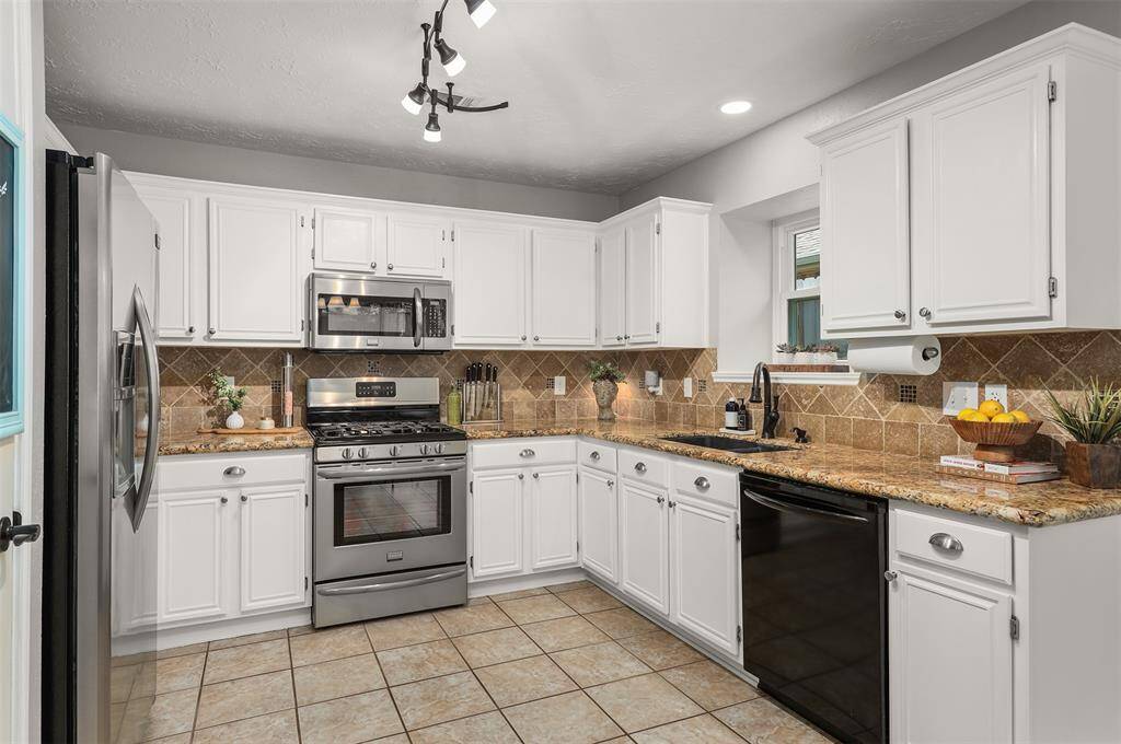 This kitchen features a modern stainless-steel range and microwave, white cabinetry, and granite countertops with a tile backsplash. It's well-lit with ample workspace and a cozy, welcoming design. Central vacuum is located right off the kitchen.