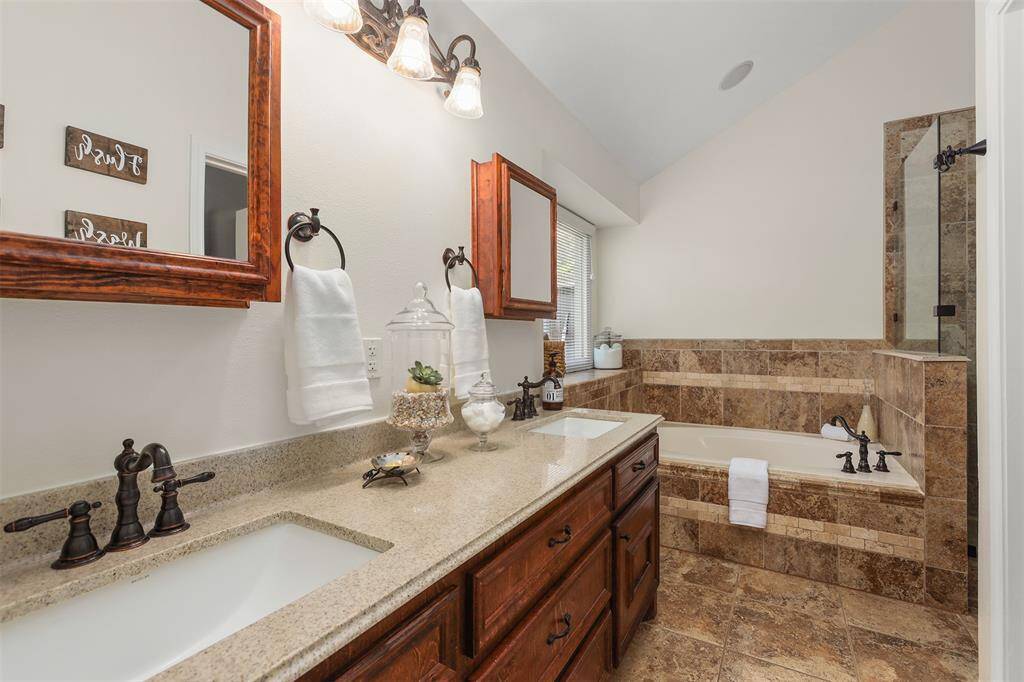 This Primary bathroom features a dual-sink vanity with granite countertops, elegant dark bronze fixtures, and separate mirrors. The space includes a luxurious tiled bathtub and a separate shower, creating a spa-like atmosphere.