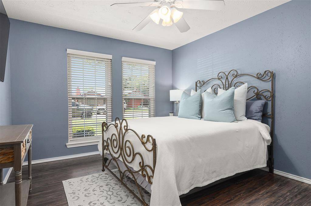 Cozy secondary bedroom with soft blue walls, ceiling fan, dark "wood like" flooring, walk in closet and large windows for natural light.