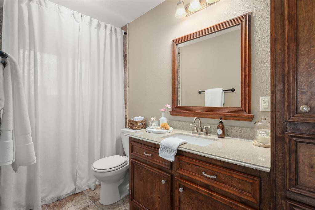 This bathroom features a classic design with a stone countertop and wooden cabinetry. It includes a large mirror, modern fixtures, and a tub/shower combo. The space is well-lit and has neutral tones, creating a warm and inviting atmosphere.