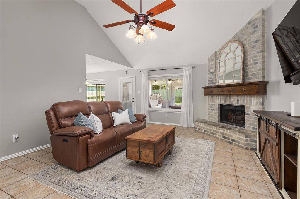 This inviting living room features a vaulted ceiling with a ceiling fan, a cozy gas log brick fireplace, and a large window looking out onto the screened in sunroom, offering plenty of natural light.