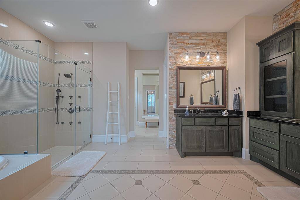 Absolute spa retreat primary bath features oversize walk-in shower, separate vanity with glass door shelving and floor to ceiling stone tile walls