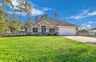 This is a single-story home featuring a two-car garage, a well-maintained lawn, and stone accents around the entrance. It's set against a backdrop of tall trees under a clear sky.