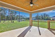 This is a spacious covered patio with a wooden ceiling and ceiling fan, offering a view of a large, fenced backyard with green grass and mature trees. Ideal for outdoor relaxation and entertainment.