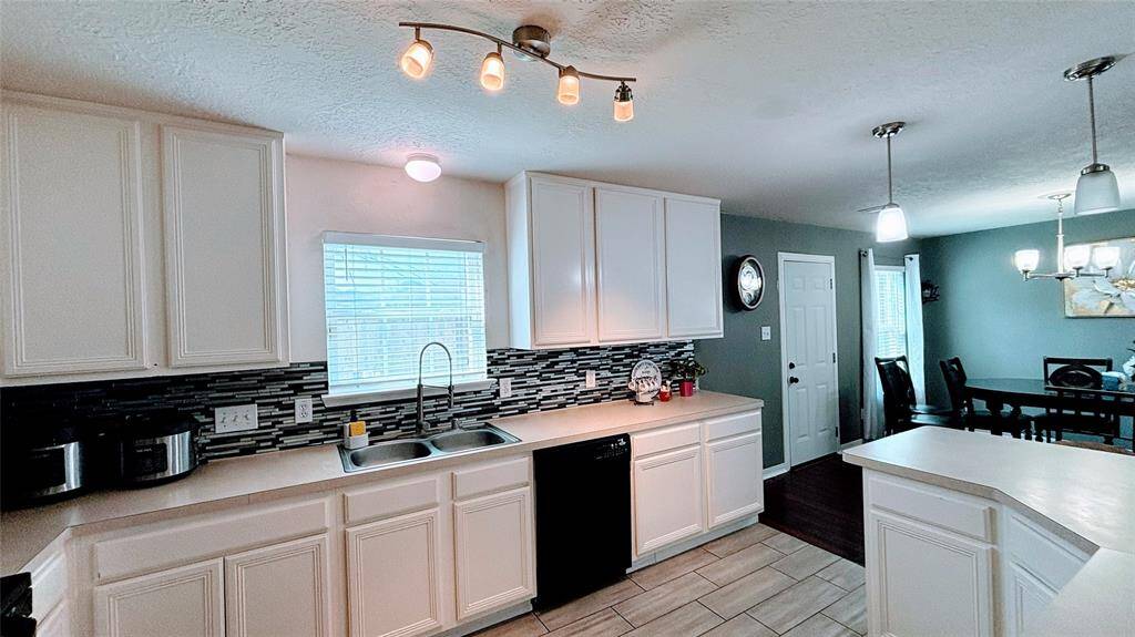 Modern kitchen with white cabinetry, a stylish tiled backsplash, and a double sink beneath a window. The kitchen features a black dishwasher and ample counter space, enhanced by contemporary lighting.