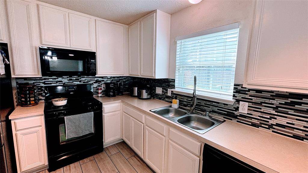 This kitchen features white cabinetry, a sleek black stove and microwave, a stylish tile backsplash, and a double sink beneath a bright window, offering ample natural light and a functional workspace.