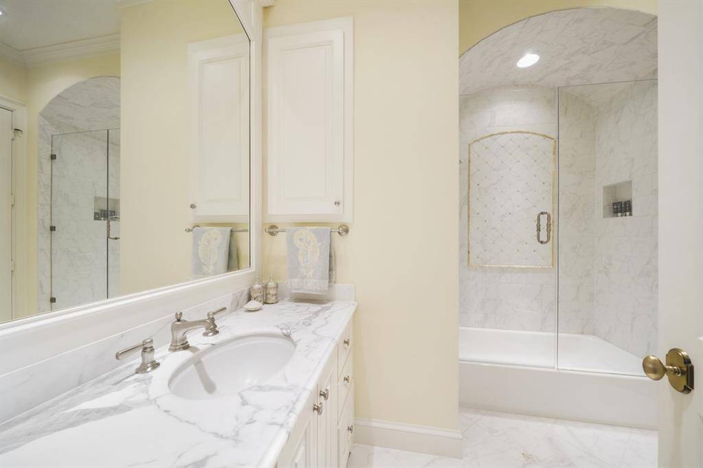 En suite full bath has a frameless shower/tub with Carrara Marble countertop and Honey Onyx accents on the tile floor and the shower tile. Built-in shower cubby to store bathing essentials. Arched paneled mirror, built-in medicine cabinet, and Kohler fixtures.