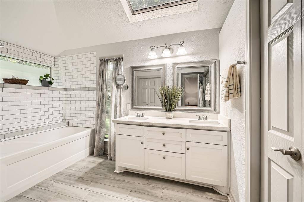 3106 Village Springs Ct. This bathroom features a modern design with a skylight, dual mirrors, and a spacious vanity. It includes a white subway-tiled bathtub, light wood flooring, and ample natural light, creating a bright and inviting space.