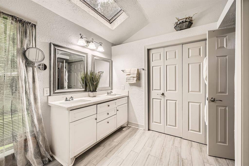 3106 Village Springs Ct. This bathroom features a modern double vanity with ample storage, two large mirrors, and a skylight that brightens the space. Neutral tones and wood-like tile flooring create an inviting atmosphere, complemented by a textured curtain and closet for added convenience.