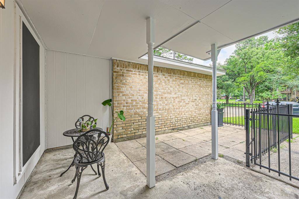 3106 Village Springs CT shows a cozy, covered patio with a small table and chairs, perfect for outdoor relaxation. It features a brick wall, concrete flooring, and a view of a green, tree-lined street through a black metal fence.