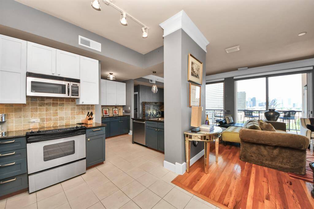 This modern open-concept apartment features a stylish kitchen with two-tone cabinets and stainless steel appliances. The living area boasts hardwood floors, abundant natural light from large windows, and access to a spacious balcony with city views.