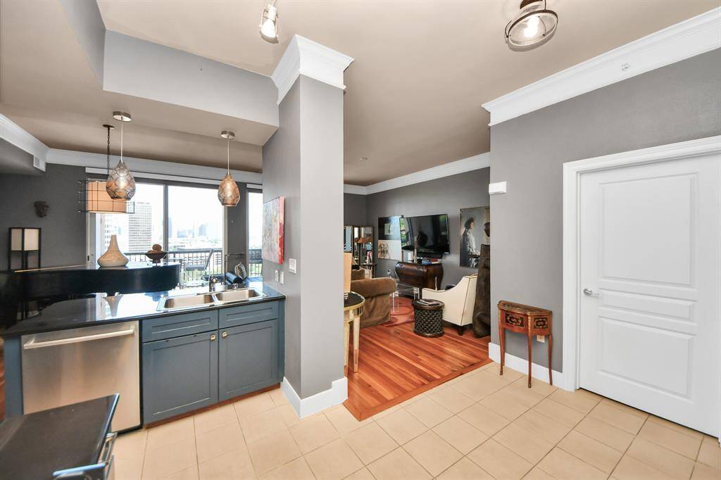 This photo showcases a modern, open-concept living space with a kitchen featuring dark cabinetry, stainless steel appliances, and pendant lighting. The adjacent living area has warm wood flooring, a large TV, and ample natural light from large windows and a balcony view.