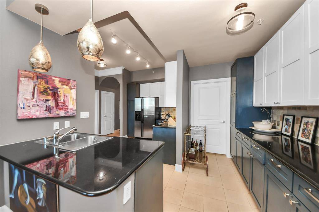 This modern kitchen features sleek black countertops, white and blue cabinetry, and stainless steel appliances. It includes stylish pendant lighting, a double sink, and a tile floor. The open layout provides ample storage and a contemporary feel, enhanced by vibrant artwork.