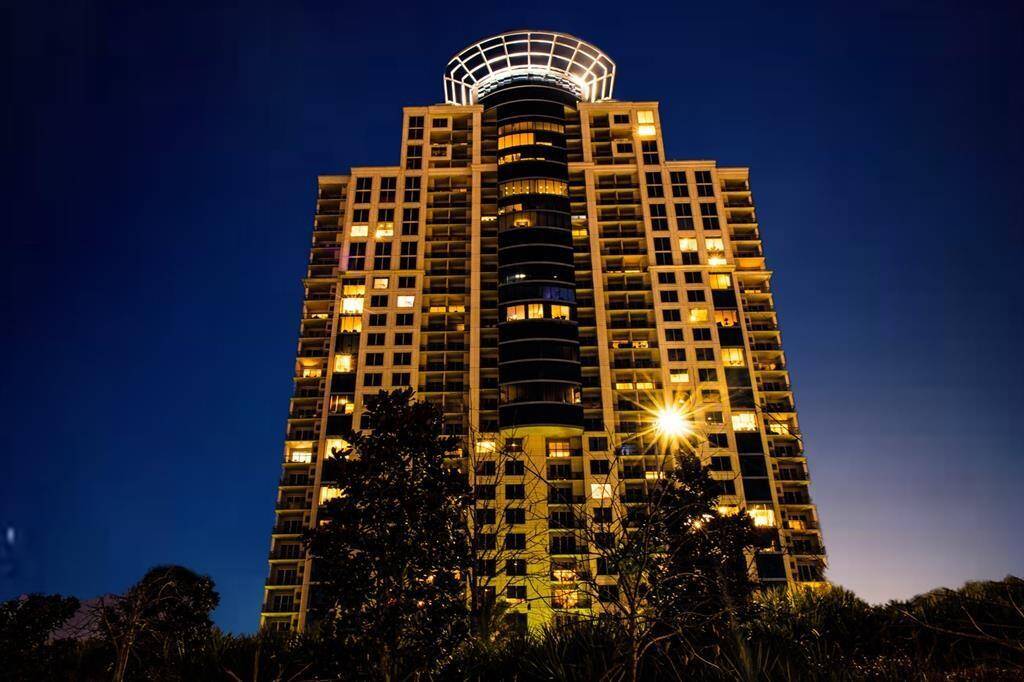 This photo showcases a modern high-rise apartment building at night, with numerous illuminated windows hinting at vibrant city living. The architectural design features a distinctive circular structure at the top, and the surrounding trees add a touch of greenery.