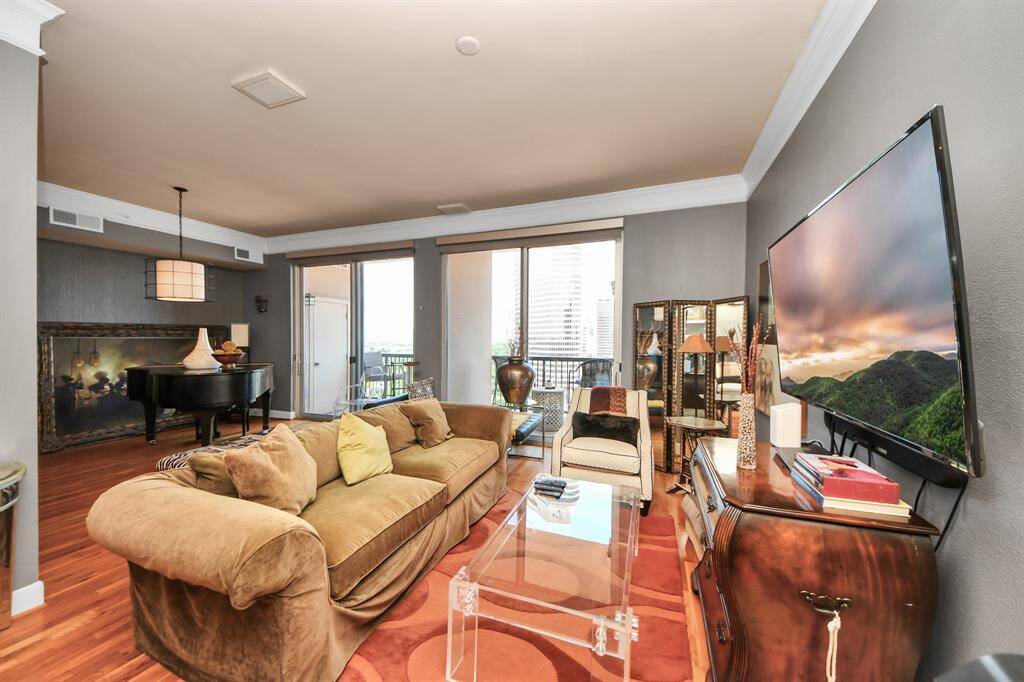 This spacious living room features warm hardwood floors, a cozy sofa, and a large wall-mounted TV. It opens to a balcony with city views, offering a bright and inviting atmosphere. A grand piano and tasteful decor add elegance to the space.