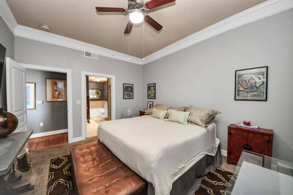 This bedroom features neutral walls, crown molding, and a ceiling fan. It includes a large bed with a padded bench at the foot, and is connected to a bathroom. The decor includes modern artwork and a red side table, offering a comfortable and stylish retreat.