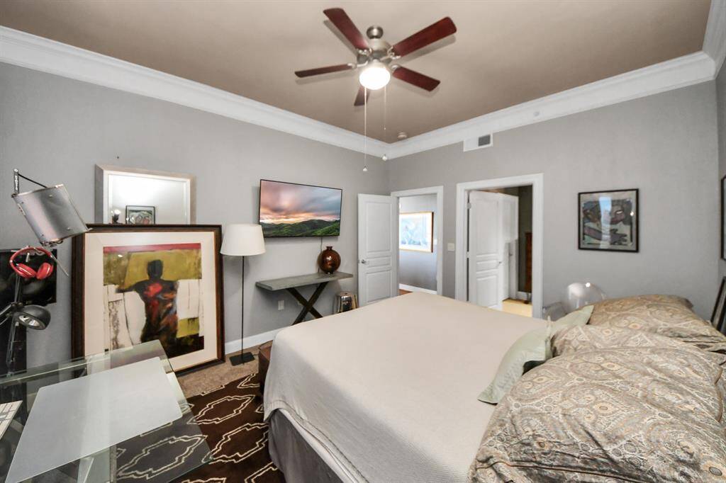 This bedroom features neutral gray walls, a ceiling fan, and modern decor. It includes a large bed, artwork, and a small desk, providing a cozy and functional space.