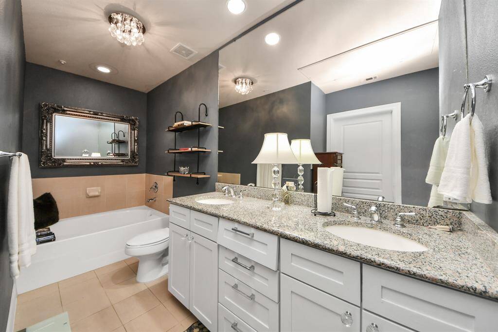 This bathroom features a modern design with a granite double vanity, elegant lighting, and a large mirror. It includes a bathtub with stylish shelving and a neutral color palette for a sophisticated look.
