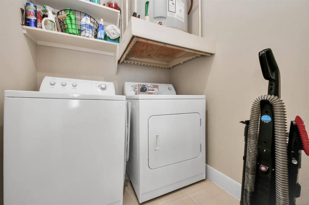 This photo shows a compact laundry area featuring a washer and dryer. Above are shelves stocked with cleaning supplies, and there's a water heater. A vacuum cleaner is positioned nearby, highlighting the space's functionality and storage options.