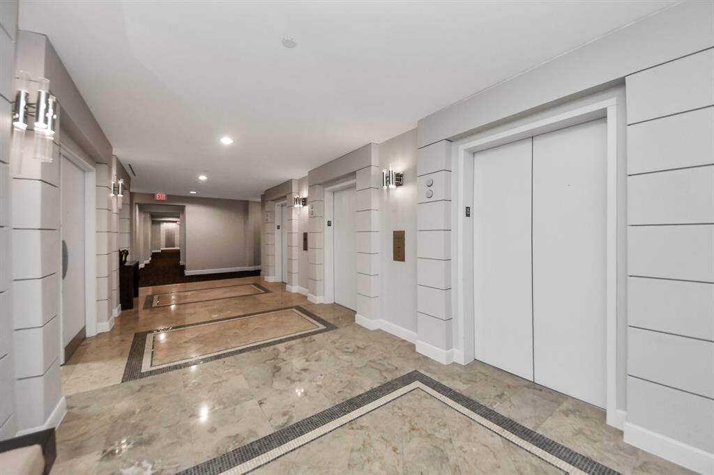 This is a modern building hallway with multiple elevators. It features sleek tile flooring, soft overhead lighting, and contemporary wall sconces, creating an inviting and well-maintained atmosphere.