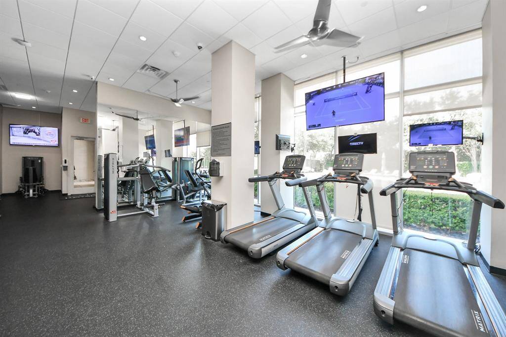 Bright and modern gym with large windows, equipped with treadmills, exercise machines, and multiple wall-mounted TVs for entertainment.